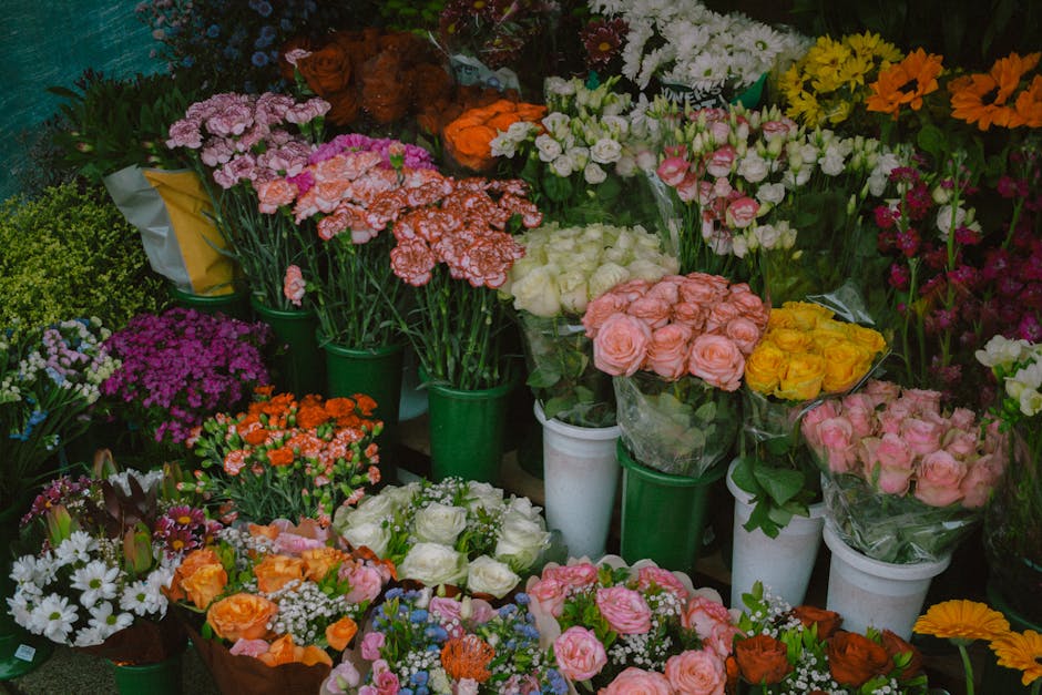 A vibrant display of fresh flower bouquets and potted plants arranged on shelves and tables, featuring a variety of blooms including pink roses, white roses, orange and yellow carnations, purple and w