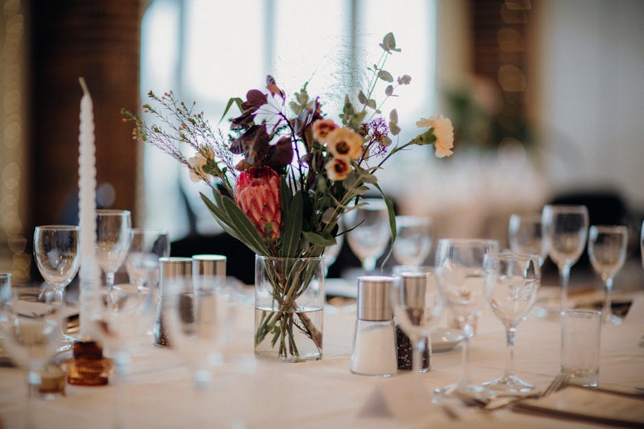 A floral arrangement featuring a variety of fresh flowers in a clear glass vase, placed centrally on a dining table set for an event at a venue with soft, natural lighting. The bouquet includes pink a