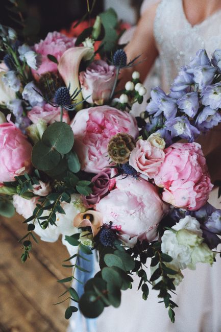 A close-up of a bridal bouquet featuring large, soft pink peonies with ruffled petals, complemented by delicate light purple and lavender flowers, possibly sweet peas or irises. The arrangement includ