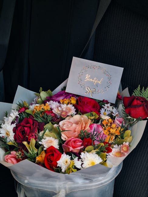 A vibrant floral arrangement featuring a mix of roses in deep red, soft pink, and peach, along with white daisies, small yellow and orange blooms, and green foliage. The bouquet is wrapped in white ti