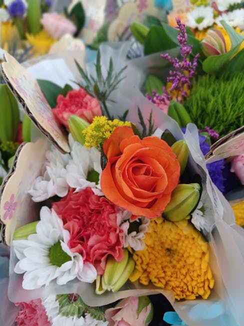 A vibrant floral arrangement featuring a large orange rose at the center, surrounded by white daisies, pink carnations, yellow chrysanthemums, and purple statice, all wrapped in decorative paper. The 