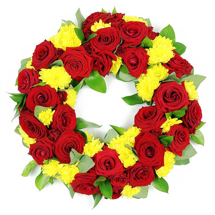 A beautifully composed funeral wreath, photographed against a stark white background, showcases a complete circle of flowers in vivid red and yellow tones, carefully crafted to convey respect and remembrance. The open-centred ring is densely filled so that each bloom touches the next, creating a continuous band of colour with no visible gaps, much like a garland of petals shaped by hand in a Muswell Hill workshop. Dominating the wreath are numerous deep red roses, each one fully opened with layered, velvety petals that create a classic spiral rose form. Their colours vary subtly from bright, romantic crimson to deeper shades approaching claret, which gives the arrangement a rich, almost ceremonial gravity. Nestled among and around these roses are clusters of yellow chrysanthemums, likely spray or button varieties, with petal-packed, cushion-like heads. These chrysanthemums introduce a soft, almost fluffy texture that contrasts gently with the sleek rose petals, while their warm golden yellow tones bring a sense of light, similar to the afternoon sun falling across the slopes near Highgate Wood. Framing the blooms is an assortment of lush greenery, including broad, dark green leaves with a gentle shine and some paler, more silvery foliage resembling eucalyptus. The greens serve both as structure and subtle decoration, adding shape, depth, and a hint of woodland freshness. The clean, bright lighting in the image highlights every contour and texture, underlining the skill that went into balancing the colours and shapes. Suitable for a service at a local Muswell Hill church or crematorium, this wreath feels like a thoughtful way to say farewell, combining the love symbolised by red roses with the comforting, hopeful message of yellow chrysanthemums.